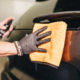 A detailer wearing black,protective detailing gloves cleans up wax residue from a car surface using an orange ADBL Puffy Towel.